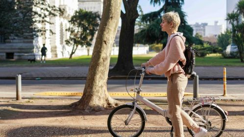 young woman walking down the street of the city. She is carrying a backpack on her back and bicycle with her hands. Profile view. Horizontal plane. Copy space for text on left side.